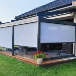 Pergola tarasowa ze zwiniętym dachem tkaninowym i z bocznymi roletami typu refleksole ZIP L wykonanymi z półprzezroczystej tkaniny, zamontowana przy nowoczesnym domu jednorodzinnym. Pod zadaszeniem znajduje się zestaw mebli ogrodowych, a wokół ustawiono donice z kwiatami, po prawej stronie pergoli zestaw do grillowania. Taras otacza zadbany zielony trawnik.