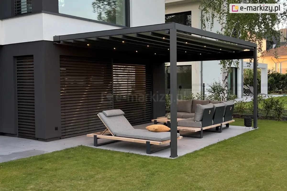 Pergola tarasowa Square Flat o szerokości 5 metrów z rozciągniętym dachem z tkaniny i zintegrowanym oświetleniem punktowym LED, zamontowana przy ciemnej elewacji nowoczesnego domu z żaluzjami fasadowymi, osłaniająca narożną sofę i leżak na tarasie ogrodowym z płyt gresowych, otoczona trawnikiem i ogrodem z brzozami.
