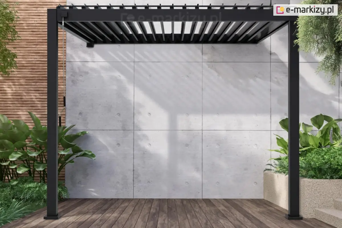 Mała pergola bioklimatyczna Gloria z obracanymi lamelami w kolorze antracytowym, montowana przy ścianie budynku. Konstrukcja wykonana z trwałego aluminium, wyposażona w ręczną korbkę do precyzyjnego sterowania kątem nachylenia lameli dachowych. Otoczenie stanowią donice z roślinami i betonowa ściana, co podkreśla nowoczesny charakter przestrzeni.