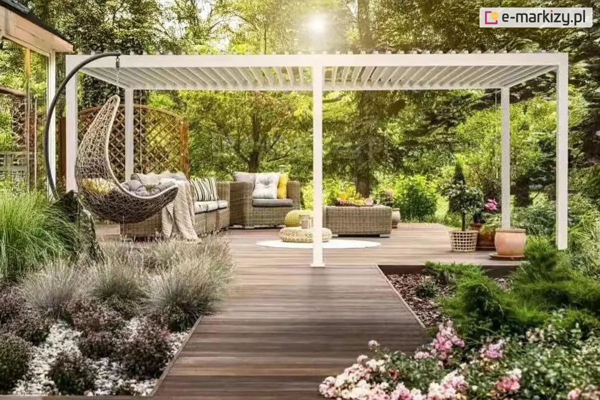 Duża biała pergola tarasowa Gloria z aluminiową konstrukcją i regulowanymi lamelami dachowymi, zamontowana w przydomowym ogrodzie z zadbaną roślinnością. W strefie relaksu ustawione są nowoczesne meble z technorattanu, fotele, poduszki i huśtawka wisząca typu kokon. Idealna osłona przeciwsłoneczna i przeciwdeszczowa na taras w stylu boho lub nowoczesnym.