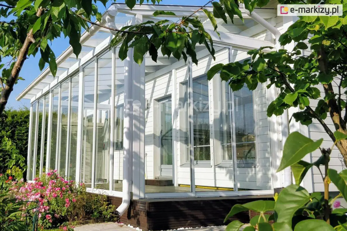 Zabudowa tarasu Sotro z białą aluminiową konstrukcją i dużymi panelami szklanymi wkomponowane w otaczający ogród przy białym domku.