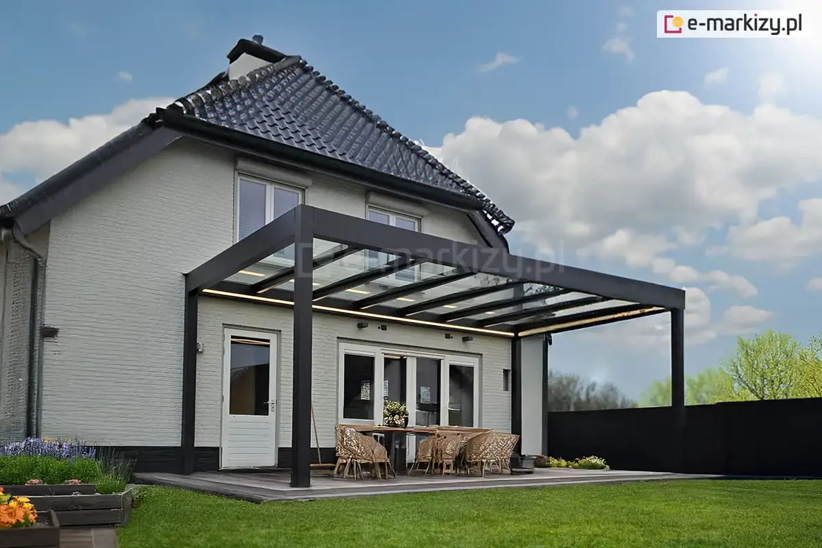 Wypoczynkowa część tarasu ze szklanym zadaszeniem Spekta Glas w kolorze antracytowym postawionym przy białym ceglanym domu z obszernym trawnikiem i kwiatami w drewnianych donicach