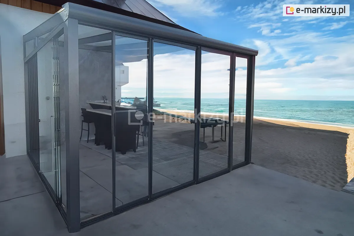 Eleganckie bistro na plaży osłonięte przesuwnym systemem drzwi szklanych Sotro w antracytowej ramie aluminiowej. Jasna, słoneczna aura i błękitne niebo podkreślają wakacyjny charakter miejsca, zapewniając komfort gościom oraz ochronę przed wiatrem, przy jednoczesnym zachowaniu malowniczego widoku na morze.