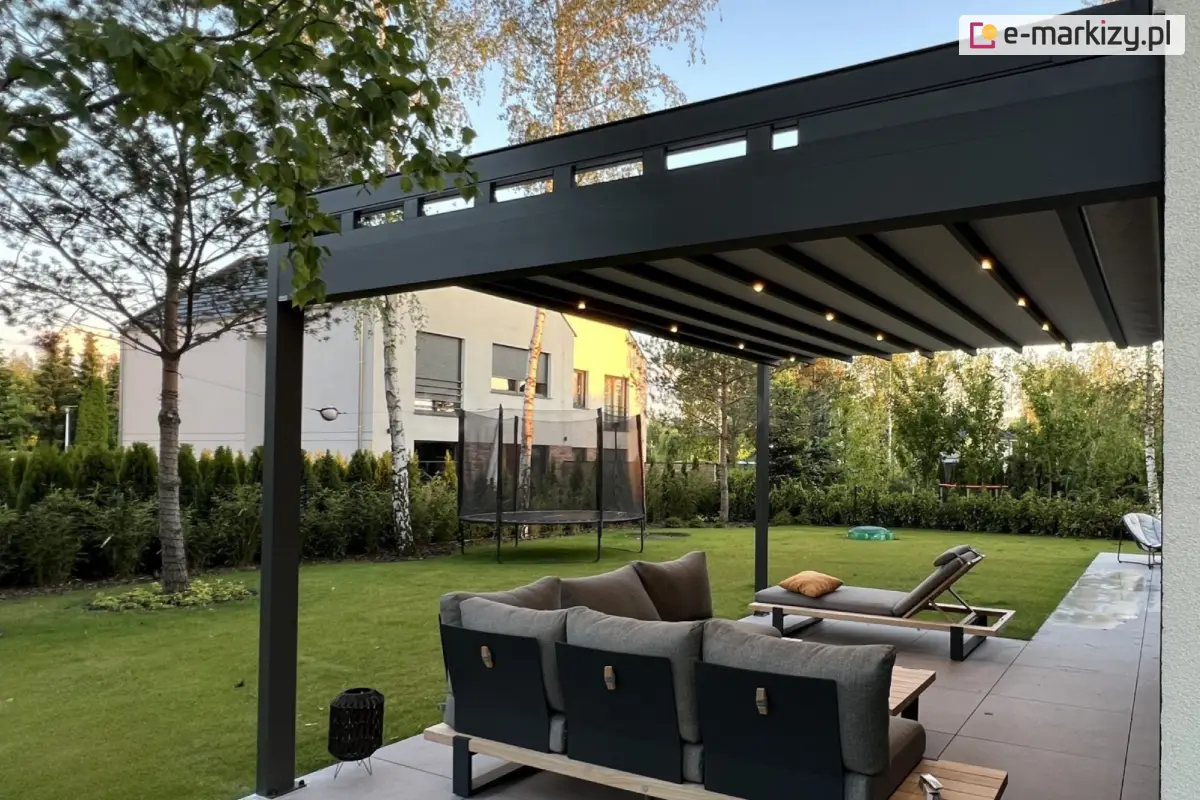 Aluminiowa pergola tarasowa Square Flat z boczną rynną odpływową po prawej stronie, z zamkniętym dachem z tkaniny i punktowym oświetleniem LED, zamontowana przy jasnej elewacji nowoczesnego domu z widokiem na zadbany ogród, trampolinę i żywopłot z tuj. Na tarasie narożna sofa i leżak z poduszkami na betonowych płytach.