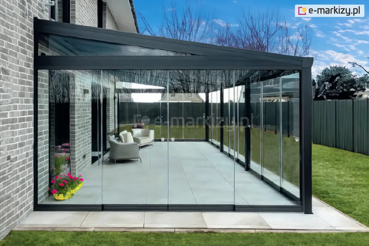 Boki grafitowej pergoli zabudowane szybami bezramowymi Miros w systemie całoszklanym, tworząc nowoczesną i elegancką przestrzeń z meblami tarasowymi chronioną przed wiatrem