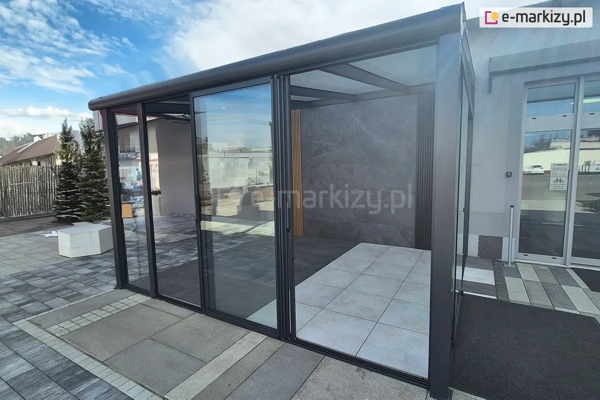 ściany przesuwne ramowe Sotro – Galeria - Całoroczny, nowoczesny ogród zimowy wyposażony w przesuwne drzwi szklane Sotro, osadzone w eleganckiej, grafitowej ramie aluminiowej. Zdjęcie ukazuje jasne, przestronne wnętrze z podłogą wyłożoną białymi i antracytowymi płytami tarasowymi.