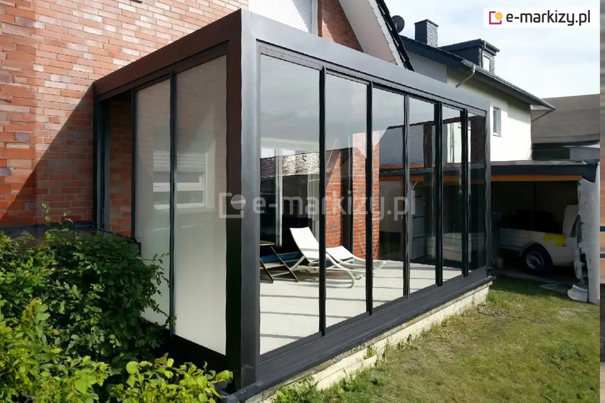 Nowoczesny czarny ogród zimowy wykonany z konstrukcji aluminiowej i przesuwnych paneli szklanych Sotro, zamontowany przy elewacji domu z czerwonej cegły. W środku widoczne białe leżaki wypoczynkowe, a całość harmonijnie komponuje się z zielenią ogrodu i sąsiednią architekturą.