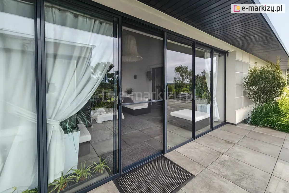 Szklane drzwi przesuwne Sotro z czarną ramą aluminiową i wewnętrznymi białymi zasłonami na tarasie nowoczesnego domu z jasnymi meblami tarasowymi