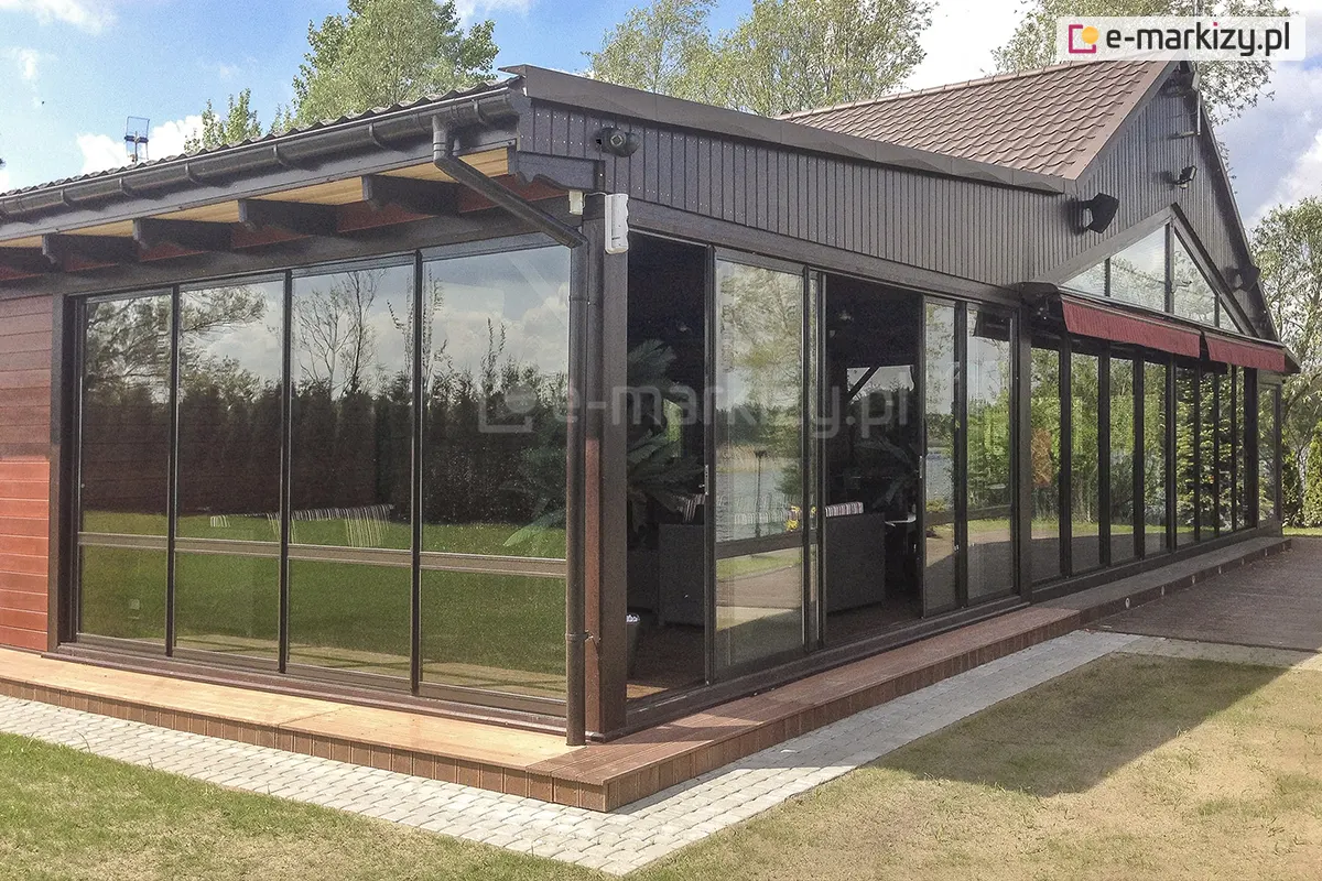 Przestronny taras zabudowany szklanym systemem ramowym Sotro z wieloma niezależnymi panelami przesuwnymi. Konstrukcja aluminiowa w ciemnym kolorze zintegrowana z budynkiem mieszkalnym, otoczona zielonym trawnikiem i ogrodem.