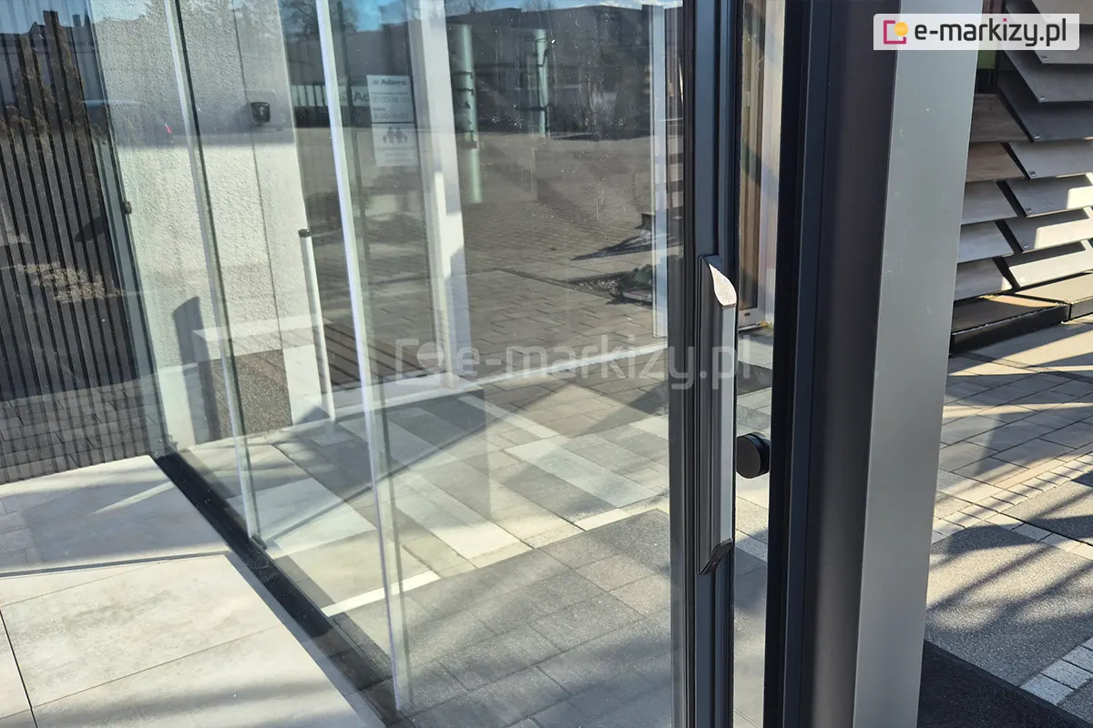 Przejrzyste szyby na bocznej ścianie pergoli z elegancką klamką zamontowaną na aluminiowej ramie przeszklonego panelu systemu Sotro.