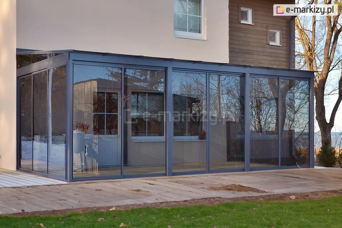 Nowoczesny ganek przy domu wykonany z aluminiowej konstrukcji oraz szklanych drzwi przesuwnych systemu Sotro z antracytową ramą. Konstrukcja w kolorze grafitowym z dużymi przeszklonymi powierzchniami, otoczona drewnianym tarasem i ogrodem.