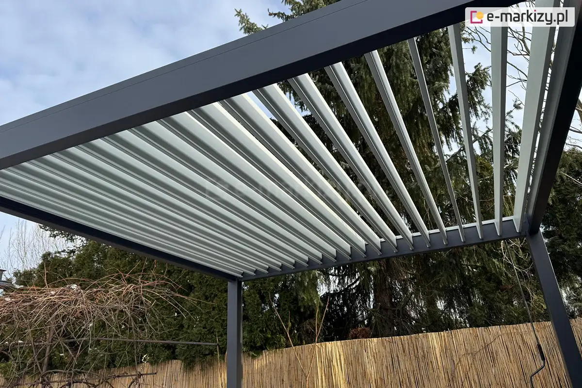 Aluminiowa pergola ogrodowa Marlo z ruchomymi lamelami dachowymi w pozycji otwartej, umożliwiająca regulację nasłonecznienia. Konstrukcja w kolorze antracytowym z jasnoszarym dachem osadzona w przydomowym ogrodzie z bambusowym ogrodzeniem i drzewami w tle. Idealna do nowoczesnych aranżacji ogrodowych.