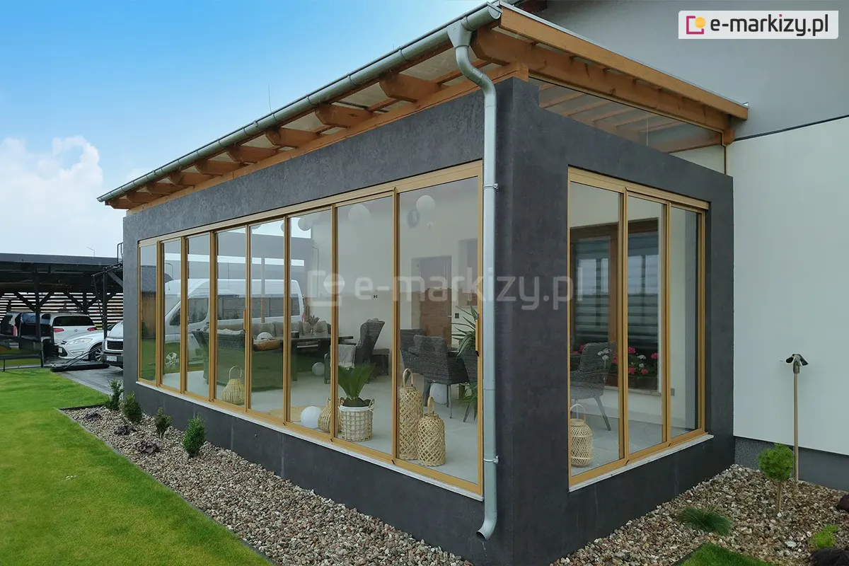 Nowoczesny ocieplany taras przy domu, zabudowany systemem szklanych drzwi przesuwnych Sotro z ramami w kolorze jasnego drewna. Konstrukcja wbudowana w antracytową ścianę elewacyjną, z zadaszeniem z drewna oraz szklanym pokryciem.