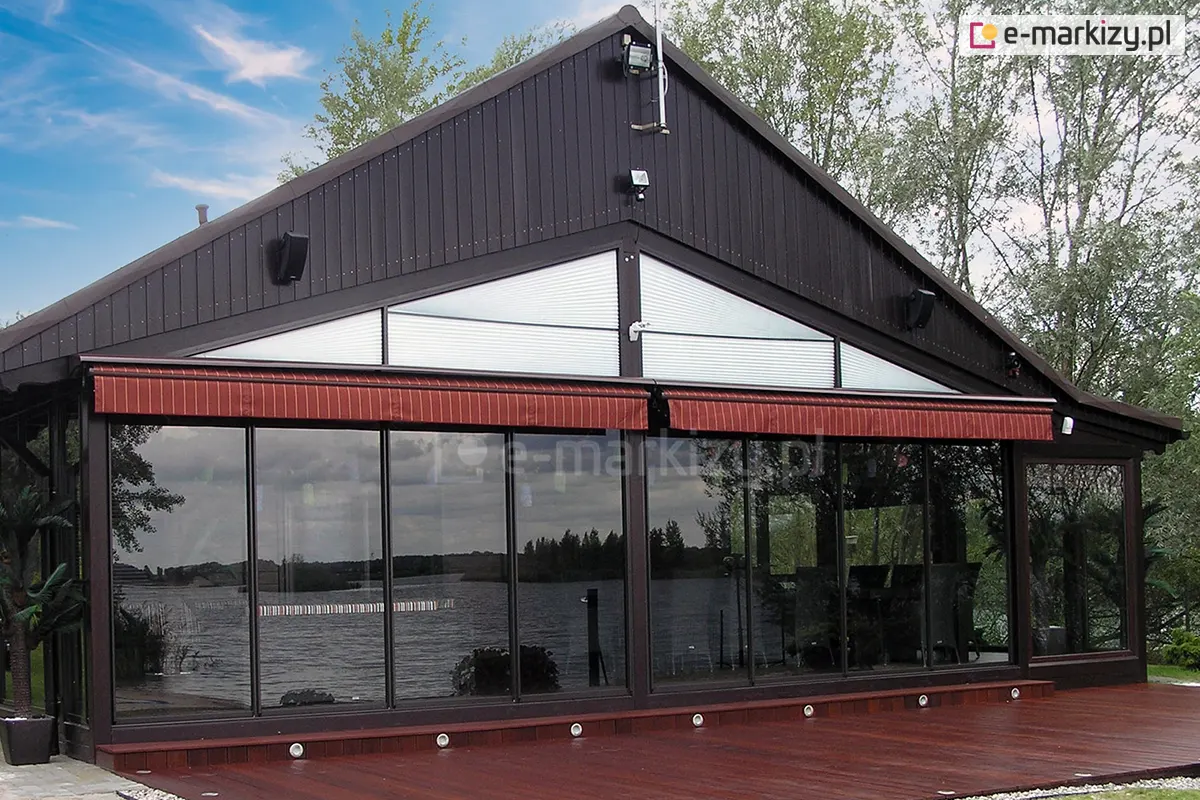 Widok na ogród letni zabudowany szklanym systemem przesuwnym Sotro przy restauracji, z czarną konstrukcją aluminiową oraz dwoma czerwonymi markizami zamontowanymi nad szybami.