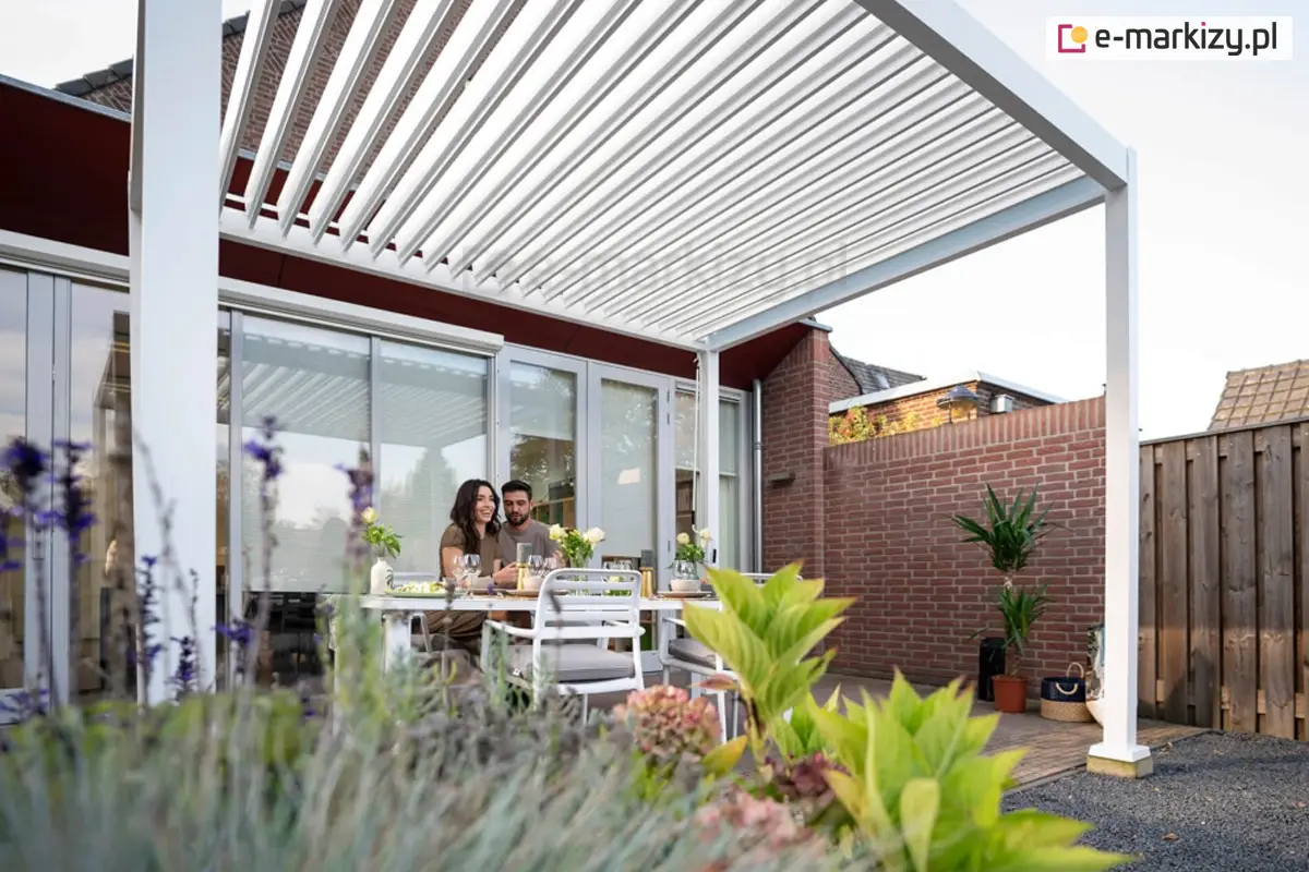 Nowoczesna aluminiowa pergola bioklimatyczna Gloria w kolorze białym zamontowana przy tarasie domu jednorodzinnego. Para siedzi przy stole pod otwartym dachem z obrotowymi lamelami, ciesząc się komfortem cienia i wentylacji. Przestrzeń tarasowa zaaranżowana w stylu ogrodowym z kwiatami i roślinami doniczkowymi. Idealne miejsce do wypoczynku i spożywania posiłków na świeżym powietrzu.