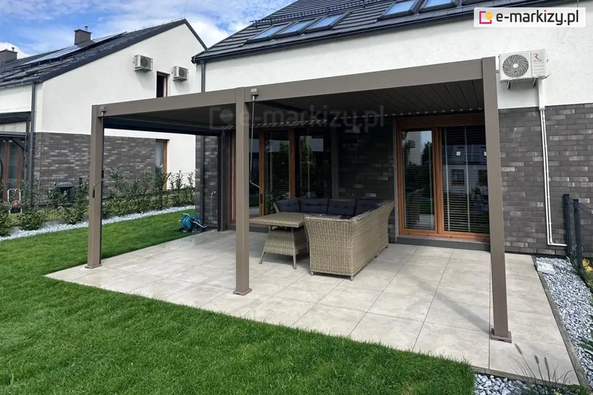 Nowoczesna pergola aluminiowa Marlo o długości 6 metrów, zamontowana przy domu jednorodzinnym z elewacją z ciemnej cegły. Konstrukcja w odcieniu brązu osłania przestrzeń wypoczynkową z technorattanowym narożnikiem i stołem. Taras wyłożony jasnymi płytkami, otoczony trawnikiem i rabatą z ozdobnymi kamieniami.