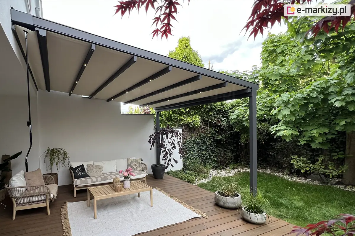 Stylowa pergola ogrodowa Aura z podświetleniem LED i wygodną korbą do sterowania tkaniną z lewej strony, osłaniająca kanapę, fotel i stolik na drewnianym tarasie w otoczeniu zieleni