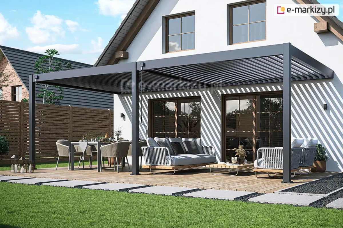 Pergola ochronna na taras Rubio Soft z aluminiowym dachem lamelowym tworzy szerokie, bioklimatyczne zadaszenie ogrodu, łączące część wypoczynkową i jadalnianą pod jednym daszkiem. Pod pergolą znajdują się nowoczesne meble ogrodowe ustawione na drewnianym podeście, a całość otacza trawnik oraz pełne prywatności ogrodzenie.