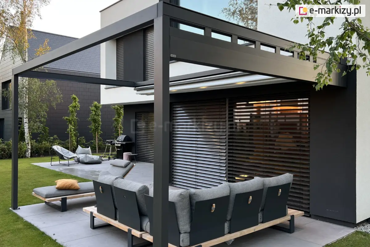 Pergola aluminiowa Square Flat z boczną rynną odprowadzającą wodę, zamontowana przy nowoczesnym domu z ciemną elewacją i dużymi oknami. Konstrukcja z aluminium w kolorze antracytowym, dach wykonany z tkaniny w pozycji częściowo zwiniętej. Pod pergolą narożna sofa ogrodowa i leżak z żółtą poduszką, w tle grill, fotele wypoczynkowe i zadbany trawnik z roślinnością.