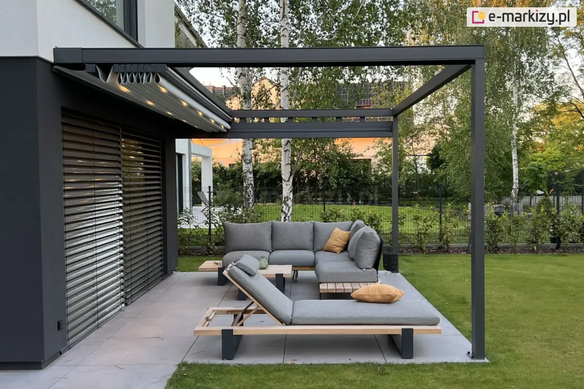 Automatyczna pergola tarasowa Square Flat z aluminiową konstrukcją w kolorze antracytowym i zwiniętą tkaniną dachową schowaną pod daszkiem, zamontowana przy elewacji domu z żaluzjami fasadowymi, wyposażona w narożną sofę i leżaki z poduszkami, ustawione na przestronnym tarasie z płyt gresowych otoczonym zielonym trawnikiem.