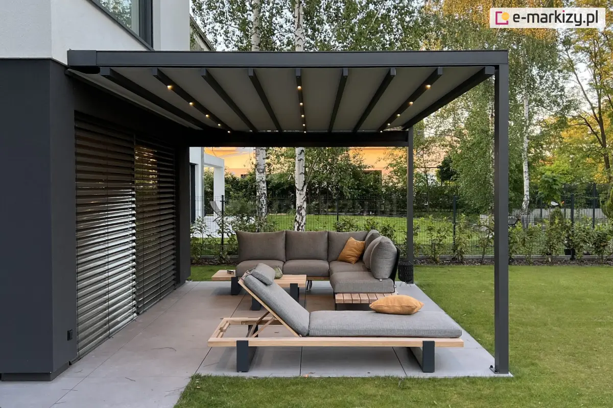 Rozwijana pergola tkaninowa Square Flat firmy Mol z oświetleniem LED w belkach dachowych i rozciągniętą tkaniną w kolorze szarym, zamontowana przy elewacji domu z żaluzjami fasadowymi, osłaniająca narożną sofę oraz leżaki z poduszkami na tarasie ogrodowym wyłożonym płytami gresowymi, otoczonym trawnikiem i zielenią.