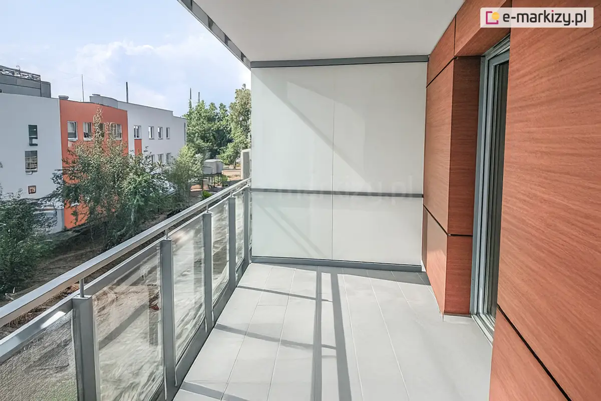Balkon w nowoczesnym bloku, z podłogą wyłożoną jasnoszarymi płytkami, ścianą wykonaną z dużych pomarańczowo-brązowych płyt oraz szklano-stalowa barierką. Pomiędzy balkonami sąsiadujących mieszkań wykonano ścianę ze stałych okien i drzwi Fest, wypełnionych mlecznym szkłem. W tle widoczny sąsiedni budynek wykonany w podobnej kolorystyce elewacji oraz drzewa przy drodze i w oddali.