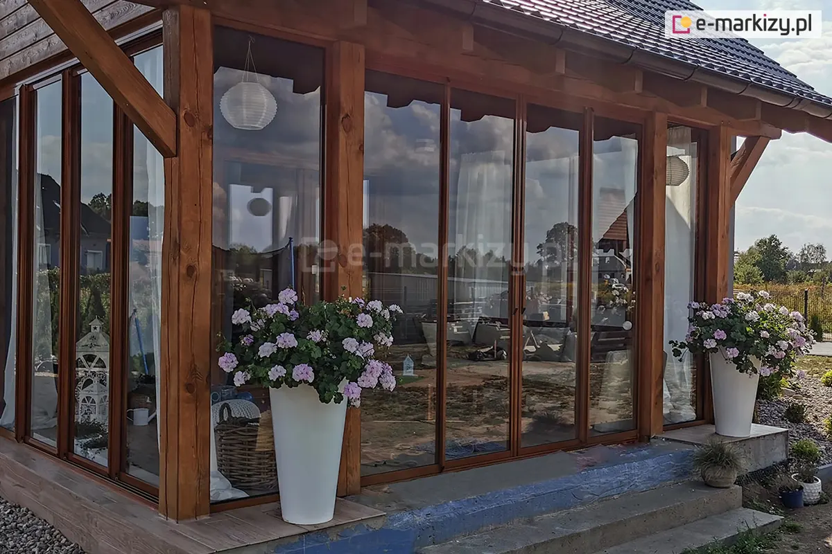 Przesuwne drzwi szklane Sotro z elegancką brązową ramą zamontowane w drewnianej konstrukcji tarasu, udekorowane kwiatami w dużych białych donicach, tworzące przytulną przestrzeń ogrodową.