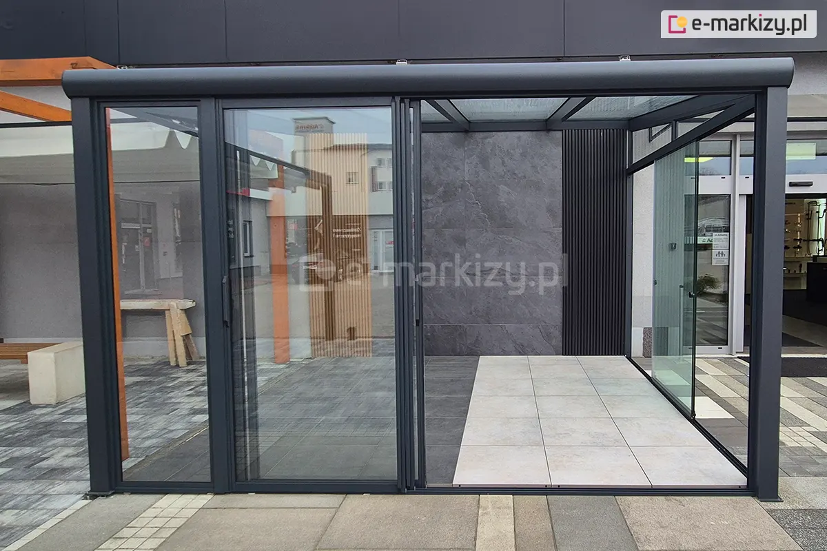 Przesuwne panele szklane Sotro z ramą aluminiową, otwarte i przesunięte na lewą stronę, prezentujące wygodny sposób dostępu do pergoli. W tle widoczne nowoczesne wnętrze pergoli o neutralnych barwach – odcieniach szarości i beżu, które tworzą elegancki, harmonijny nastrój.