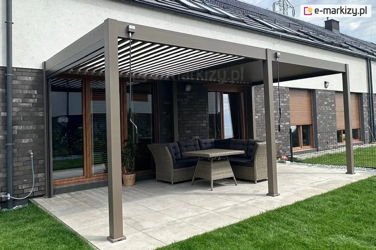 Nowoczesna pergola aluminiowa Marlo zamontowana na przydomowym tarasie, bezpośrednio przy elewacji budynku. Konstrukcja w odcieniu szarości osłania wygodny zestaw mebli ogrodowych z technorattanu – narożną sofę i stół. Taras wyłożony jasnymi płytkami, a otoczenie stanowi zadbany trawnik.