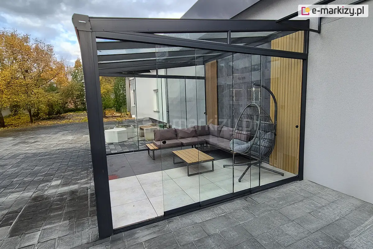Uniwersalna szklana zabudowa pergol i tarasów na wymiar Miros - przesuwne panele szklane zamontowane z każdej strony zadaszenia przy domu z jasną elewacją. Wewnątrz komfortowy zestaw wypoczynkowy z narożną sofą, stolikami i fotelem wiszącym. Otoczenie jesienne, z drzewami o złotych liściach.
