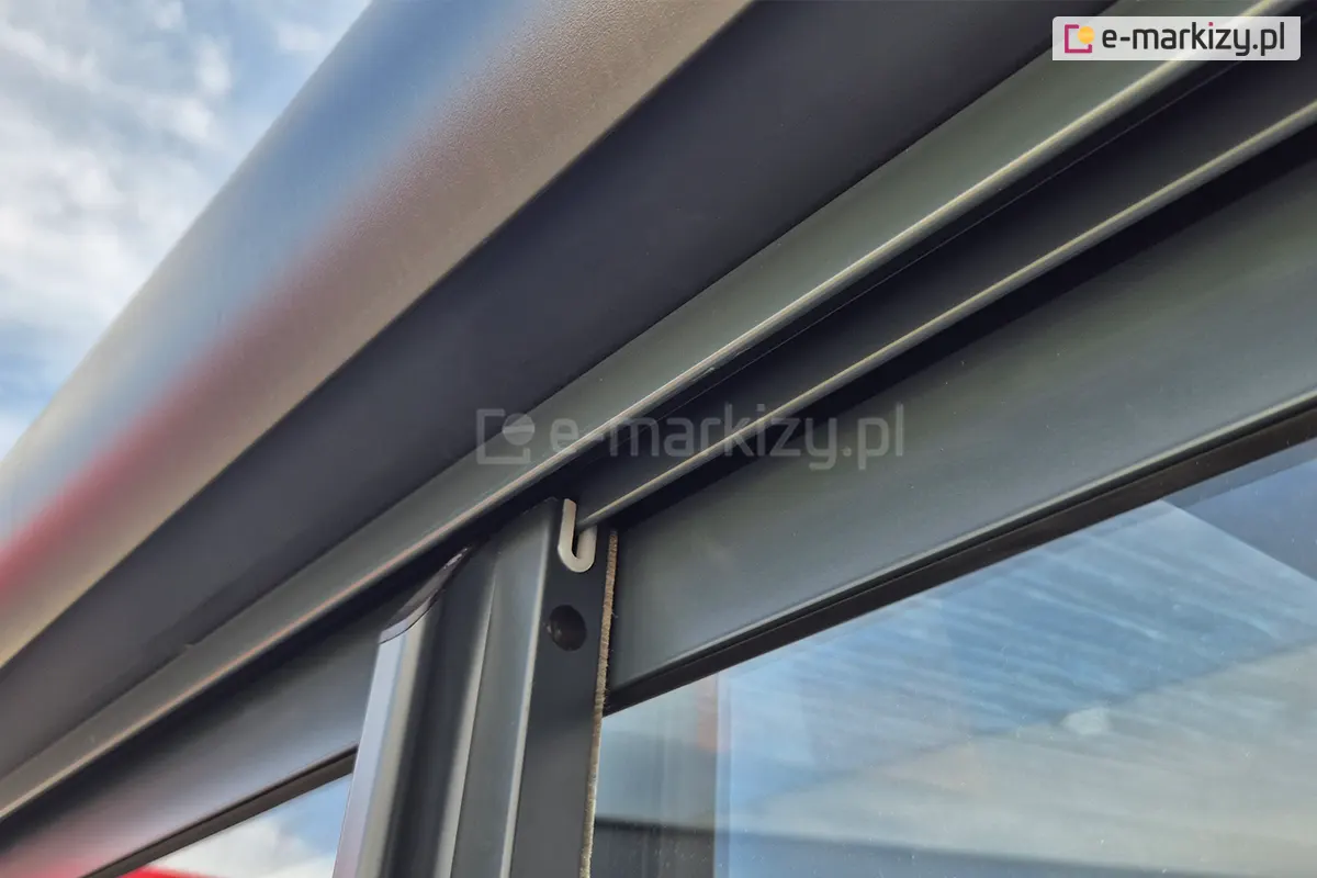 Zbliżenie na górną prowadnicę systemu Sotro, zamontowaną bezpośrednio do belki pergoli, w której osadzony jest przesuwny panel szklany w eleganckiej, ciemnoszarej ramie aluminiowej.