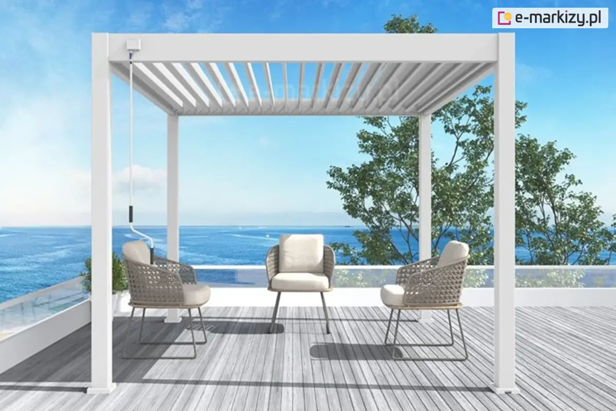 Biała pergola ogrodowa model Marlo o wymiarach 3x3 metry, ustawiona na drewnianym tarasie z widokiem na morze. Pod pergolą znajdują się trzy eleganckie fotele z jasnymi poduszkami. W tle szklana balustrada, błękitne niebo i spokojna tafla wody, tworzące relaksującą, nadmorską atmosferę.