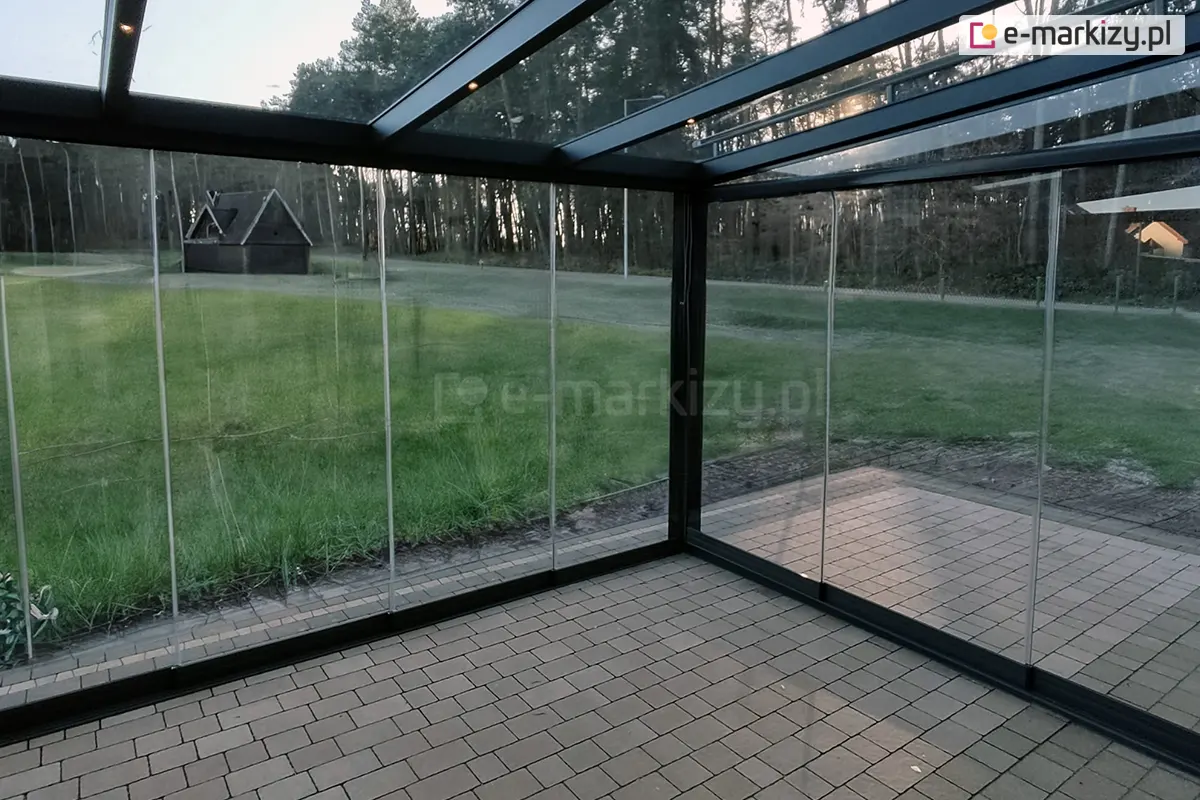 **Opis alt (SEO):** Taras zimowy zabudowany szkłem Miros z widokiem na ogród i las, prezentujący nowoczesne przesuwne panele szklane zamontowane w solidnej aluminiowej konstrukcji z przejrzystym dachem