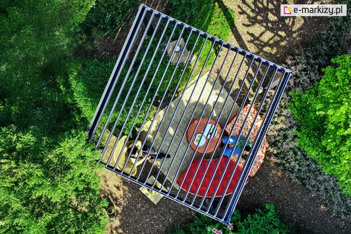 Nowoczesna pergola bioklimatyczna Gloria w wersji wolnostojącej, prezentowana z lotu ptaka, zamontowana w ogrodzie z gęstą roślinnością. Konstrukcja aluminiowa w kolorze antracytowym z dachem z regulowanych lameli zapewnia ochronę przed słońcem i deszczem. Pod pergolą znajduje się elegancka strefa relaksu z dywanem zewnętrznym, ozdobnymi pufami i stolikiem kawowym