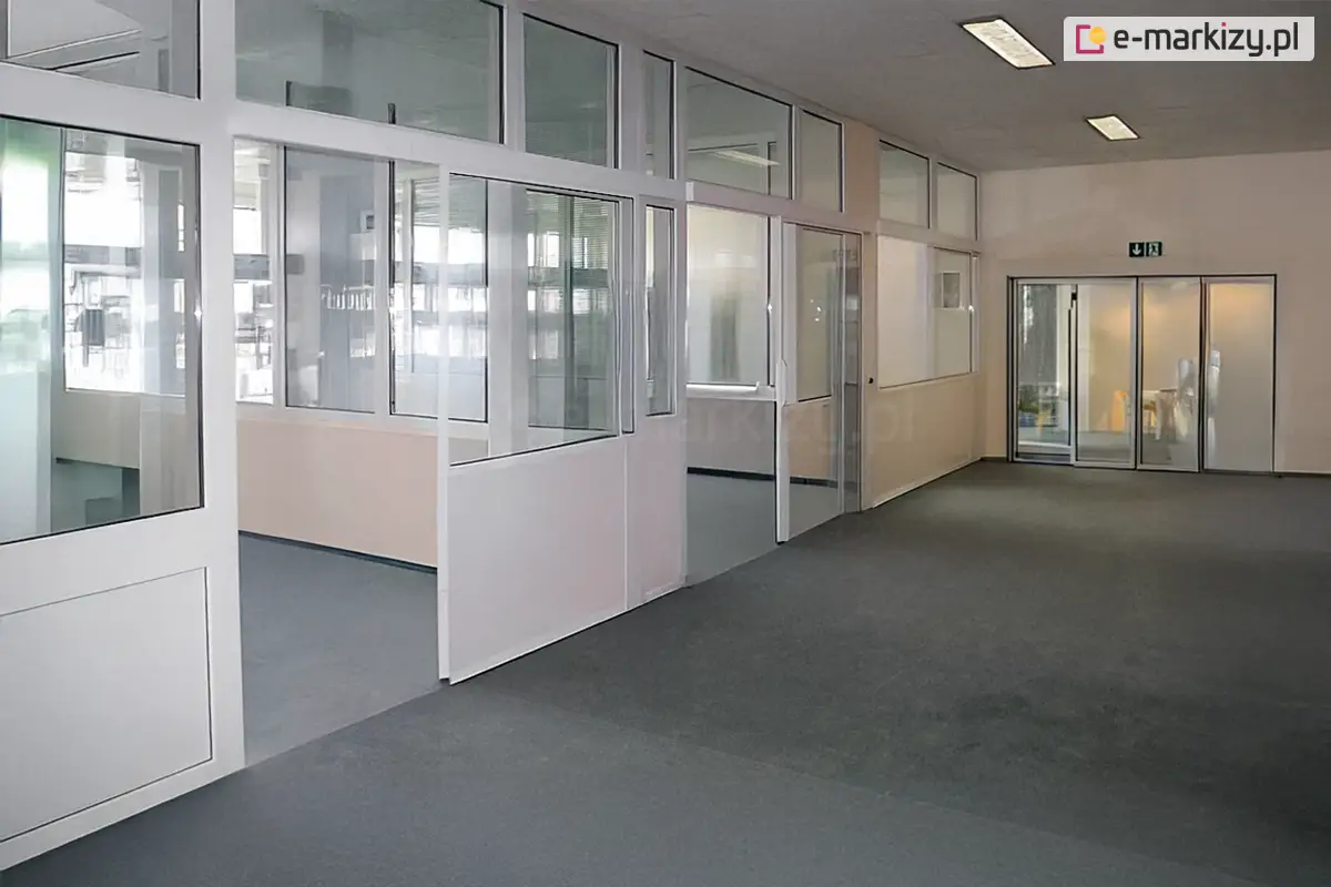 Przestronne wnętrze biurowe podzielone systemem ścianek stałych Fest, wykonanych z białych profili aluminiowych. Konstrukcja tworzy szereg przeszklonych pomieszczeń i boksów, które oddzielają strefy pracy od szerokiego ciągu komunikacyjnego. Poszczególne moduły ścianek posiadają mieszane wypełnienie: dolne sekcje wykonano z nieprzeziernych paneli w kolorze białym, natomiast górne partie są w całości przeszklone, co zapewnia swobodny przepływ światła i optyczną przestronność. W systemie zintegrowano przesuwne drzwi wejściowe do poszczególnych biur, osadzone w aluminiowych prowadnicach. Na podłodze korytarza oraz wewnątrz boksów znajduje się jednolita, ciemnoszara wykładzina obiektowa o krótkim włosiu. W głębi korytarza widoczne jest przeszklone wyjście ewakuacyjne oznaczone zielonym piktogramem, a na suficie zamontowano podłużne lampy jarzeniowe zapewniające równomierne oświetlenie techniczne. Całość aranżacji utrzymana jest w profesjonalnej, biało-szarej kolorystyce, podkreślającej sterylny i nowoczesny charakter biura.