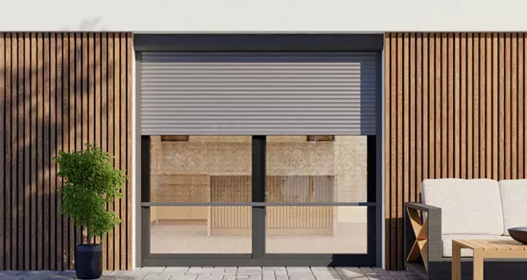 Elewacja budynku z modnymi drewnianymi lamelami oraz dużym oknem tarasowym przed którym po lewej stronie postawiono zielone drzewko w donicy, a po prawej ławkę z białymi poduchami oraz drewniany stolik. Na oknie tarasowym zamontowana roleta zewnętrzna ze skrzynką o półokrągłym kształcie oraz częściowo opuszczonym pancerzem w odcieniach szarości i wysuniętą poniżej moskitierą zintegrowaną z roletą.