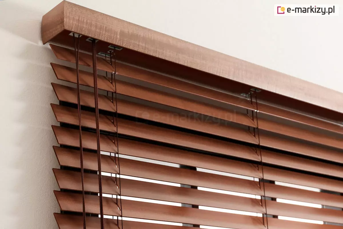 Zdjęcie prezentuje zbliżenie na górną sekcję systemu osłonowego, jakim są żaluzje drewniane 50 mm, zamontowane bezpośrednio przy jasnej ścianie. Kluczowym elementem kompozycji jest ozdobna, drewniana maskownica rynny górnej o profilowanym kształcie, wykonana z litego drewna w nasyconym, brązowym odcieniu z wyraźnym rysunkiem słojów. Pod maskownicą widoczny jest pakiet poziomo ustawionych lameli o szerokości 50 mm, które są połączone za pomocą cienkich, brązowych sznurków drabinki. W lewej części kadru znajdują się cztery pionowe sznurki operacyjne, służące do sterowania mechanizmem podnoszenia, które przechodzą przez metalowe przelotki w dolnej części rynny. Żaluzje drewniane 50 mm charakteryzują się precyzyjnym wykończeniem krawędzi oraz spójnością kolorystyczną wszystkich elementów osprzętu, co podkreśla ich elegancki i klasyczny charakter. Światło padające z boku wydobywa fakturę drewna oraz trójwymiarowość całej konstrukcji.