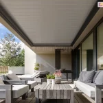 szerokie ujęcie nowoczesnego tarasu z zainstalowanym dachem lamelowym SB400R w pozycji całkowicie zamkniętej. Aluminiowe lamele w kolorze jasnoszarym tworzą szczelną, gładką płaszczyznę zadaszenia, która jest zintegrowana z ciemną ramą konstrukcyjną pergoli. Pod zadaszeniem zaaranżowano strefę wypoczynkową z meblami ogrodowymi o drewnianej konstrukcji w szarym odcieniu, wyposażonymi w grube, jasne poduszki. W centralnej części znajduje się niski stolik kawowy z surowego drewna, na którym ustawiono doniczki z wrzosami. Taras wyłożony jest deskami kompozytowymi i przylega do budynku z dużymi przeszkleniami, w których odbija się otaczająca natura. W tle widoczny jest zadbany ogród z niską roślinnością, drzewami iglastymi oraz ażurowym ogrodzeniem, a nad całością rozpościera się pogodne niebo z delikatnymi chmurami.